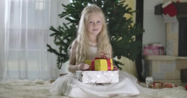 Retrato de menina branca loira bonita com olhos cinzentos em vestido branco e coroa sentado no fundo da árvore de Natal com presentes. Cinema 4k ProRes HQ . — Vídeo de Stock