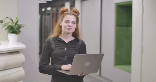 Portrait de belle rousse femme caucasienne debout avec un ordinateur portable dans un bureau ouvert, regardant la caméra et souriant. Jeune femme d'affaires prospère posant sur le lieu de travail. Siège social Cinema 4k ProRes . — Video