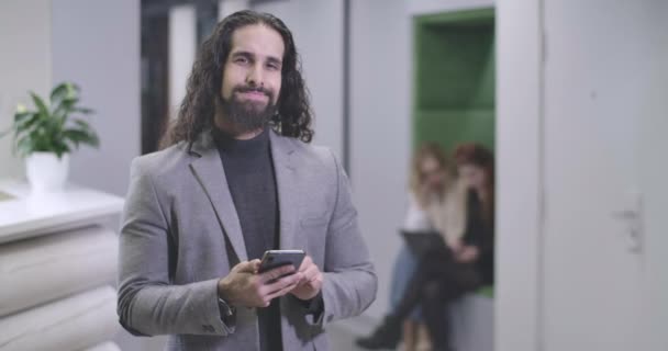 Portrait of handsome Middle Eastern man typing on smartphone keyboard, looking at camera and smiling. Confident office worker standing in open space. Business, success. Cinema 4k ProRes HQ. — Stock Video