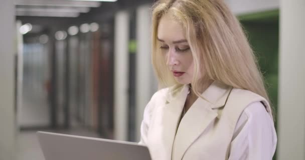 Portrait of beautiful blond Caucasian woman standing with laptop in open space office, looking at camera and smiling. Successful young businesswoman posing at workplace. Cinema 4k ProRes HQ. — Stock Video