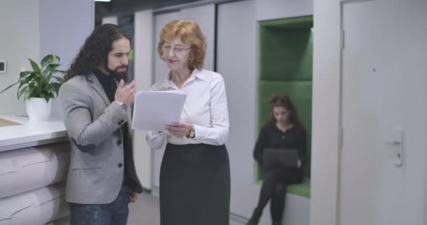 Femme caucasienne d'âge moyen en lunettes expliquant les détails du projet d'entreprise au jeune homme du Moyen-Orient. PDG aider le nouvel employé. Siège social Cinema 4k ProRes . — Video