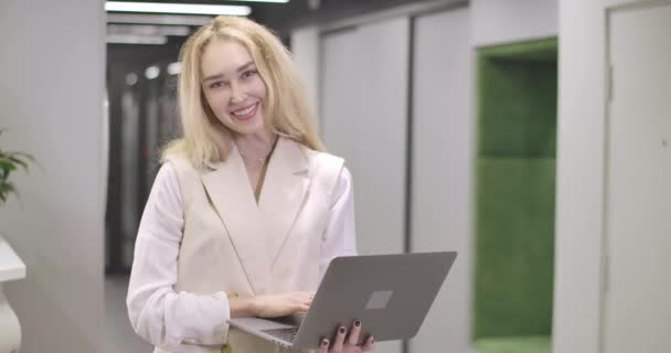 Jeune femme caucasienne attrayante debout avec ordinateur portable dans un bureau ouvert, regardant la caméra et souriant. Femme d'affaires prospère posant dans le coworking. Siège social Cinema 4k ProRes . — Video