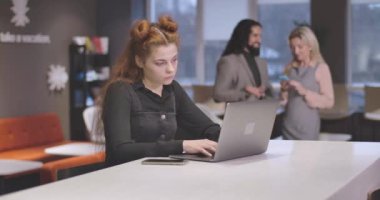 Laptop klavyesinde yazı yazan ve kafasını eliyle tutan yorgun kızıl saçlı beyaz kadın portresi. Açık uzay ofisinde aşırı çalışan yorgun bayan çalışanlar. Sinema 4k Prores Merkezi.
