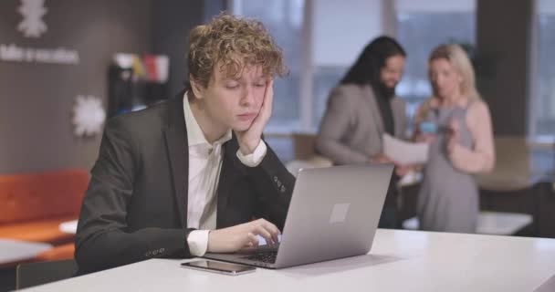 Portret van vermoeide blanke jongeman typen op laptop toetsenbord, kijken naar smartphone, en wrijven hoofd. Uitgeputte mannelijke werknemer overwerkt in open ruimte kantoor. Bioscoop 4k Prores Hq. — Stockvideo