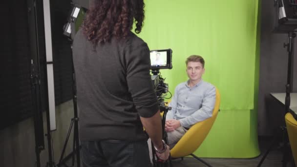 Portret van een jonge blanke man zittend op een gele fauteuil en pratend op de camera. Cameraman neemt video op. Blogger maakt materiaal klaar voor de webpagina. Vlogging, sociale media. Gericht op camerascherm. — Stockvideo