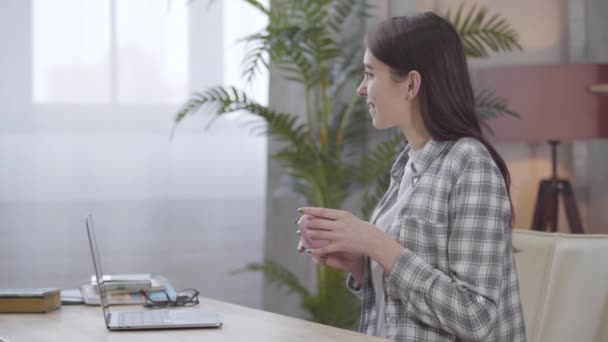 Zijaanzicht van gelukkig brunette Kaukasisch meisje zitten aan de tafel met laptop en kopje thee of koffie en glimlachen. Gelukkige freelancer die wegkijkt. Freelance, werk op afstand. — Stockvideo