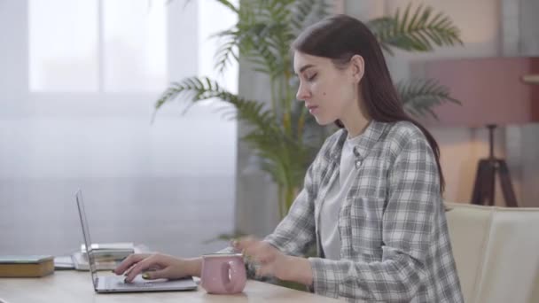 Vista lateral da jovem mulher morena caucasiana colocando corte com chá ou café na mesa e digitando no teclado do laptop. Freelancer feminino trabalhando on-line a partir de casa. Trabalho autônomo, distante . — Vídeo de Stock