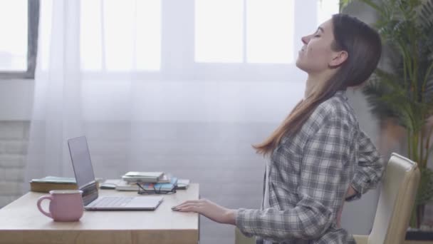 Vue latérale de la fille brune blanche confiante ayant mal au dos tout en tapant sur le clavier de l'ordinateur portable. Jeune pigiste faisant de l'exercice assis à la table. Travail à distance, position assise incorrecte . — Video