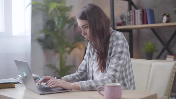 Junge brünette kaukasische Geschäftsfrau tippt auf der Laptop-Tastatur und schaut auf ihre Uhr. Freiberuflerin mit Projekttermin. Fernarbeit, freiberuflich. — Stockvideo