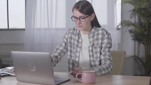 Jolie fille brune caucasienne dans des lunettes élégantes tapant sur le clavier de l'ordinateur portable. Femme indépendante souriante et confiante qui travaille en ligne depuis chez elle. Freelance, travail à distance . — Video