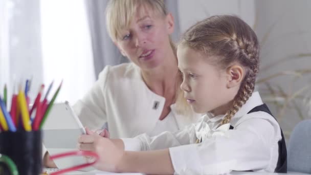 Vista lateral da linda colegial caucasiana sentada à mesa com tablet como tutor de meia-idade explicando-lhe algo no fundo. Educação, inteligência, diligência, ensino privado, tutor . — Vídeo de Stock