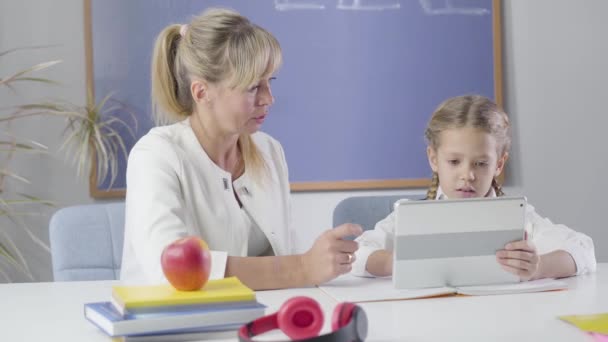 Porträtt av en vuxen vit kvinna och en liten skolflicka som sitter vid bordet med surfplatta. Smart lärare som undervisar skolflicka hemma. Utbildning, privat undervisning. Kameran rör sig från höger till vänster. — Stockvideo