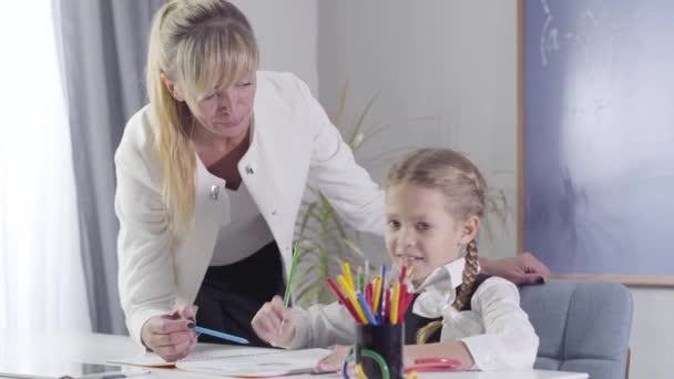 Blank schoolmeisje kiezen kleurrijke potlood om te tekenen in het oefenboek. Een kind dat thuis studeert met een leraar. Privé-onderwijs, toewijding, leren. Camera beweegt van links naar rechts. — Stockvideo