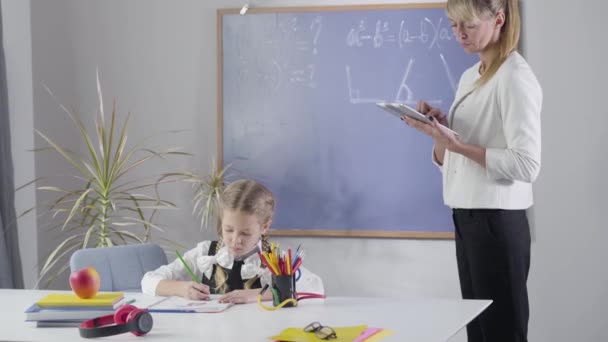 Маленькая белая девочка с косичками пишет в тетрадке, как взрослый частный учитель ищет задания в планшете. Ребенок учится дома. Частное образование, трудолюбие, обучение . — стоковое видео