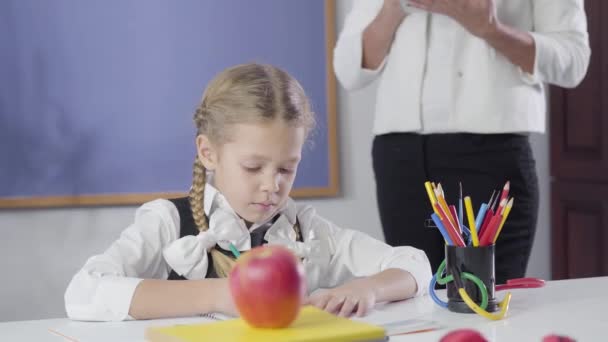 Portrait de fille caucasienne avec des nattes écrivant dans le cahier d'exercices comme professeur privé adulte debout à côté d'elle. Enfant étudiant à la maison. Enseignement privé, diligence, apprentissage . — Video