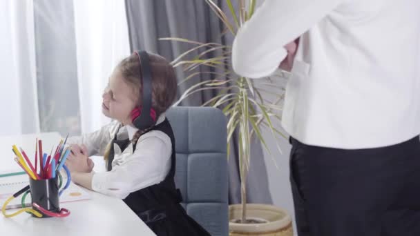Side view of cute Caucasian girl sitting in headphones and shaking head as unrecognizable female tutor standing next to her. Girl misbehaving as studying at home. Education, learning, behavior. — Stock Video