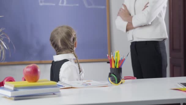 Portret małej białej uczennicy patrzącej wstecz na tablicę, odwracającej się do kamery i kładącej głowę na rękach. Niepoznawalna kobieta uczy zmęczone dziecko. Inteligencja, edukacja. — Wideo stockowe