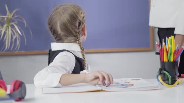 Närbild av kaukasiska skolflicka tittar tillbaka på svarta tavlan, vänder tillbaka till kameran och gör missnöjda ansikte. Oigenkännlig kvinna som lär trötta barn. Underrättelse, utbildning. — Stockvideo