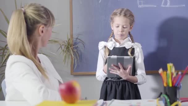 Retrato de una encantadora chica caucásica en uniforme escolar declamando a una maestra de mediana edad sentada a la mesa. Niño inteligente estudiando en casa. Enseñanza privada, tutoría, educación . — Vídeos de Stock