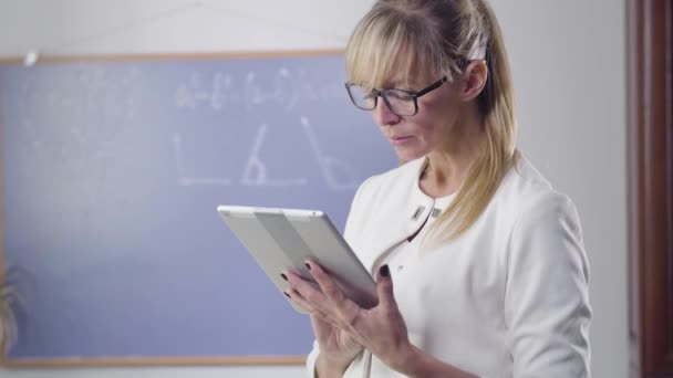 Ritratto di donna caucasica di mezza età in piedi con tablet e guardando lo schermo del dispositivo. Insegnante privato in occhiali eleganti alla ricerca di compiti interessanti. Intelligenza, istruzione, occupazione . — Video Stock