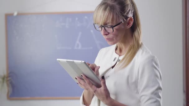 Geconcentreerde blanke vrouw van middelbare leeftijd die met een tablet staat en achterom kijkt. Privéleraar in elegante bril aan het werk. Intelligentie, onderwijs, beroep. — Stockvideo