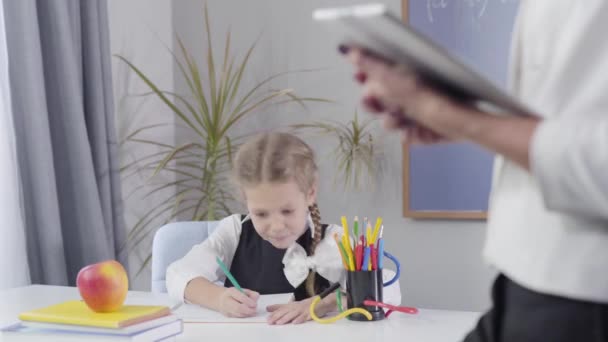 Charmig smart vit skolflicka gör jag fick en idé gest och skriva ner i träningsboken. Oigenkännlig kvinna som står i förgrunden till höger. Fokuserat på barn. — Stockvideo