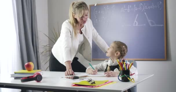 Självsäker kaukasisk medelålders lärare i formell kostym som står bredvid skolflickan och tittar på när hon skriver i träningsboken. Vuxen kvinna som undervisar flitigt barn hemma. Utbildning. Cinema 4k Prores Hq. — Stockvideo