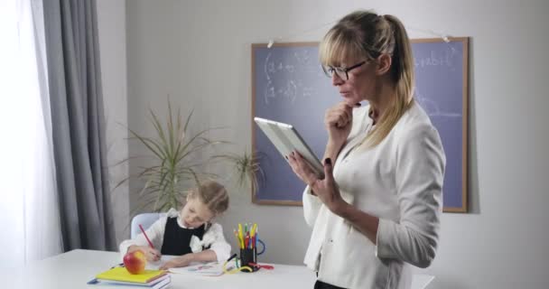 Portret van een volwassen blanke vrouw met tablet als klein mooi schoolmeisje dat in het oefenboek schrijft. Kind kijkt op en legt hoofd op handen. Intelligentie, onderwijs. Bioscoop 4k Prores Hq. — Stockvideo
