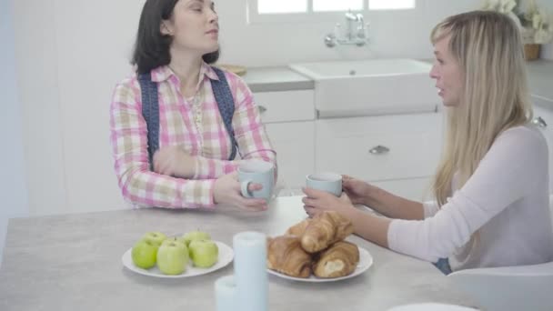 Donna caucasica adulta bionda seduta a tavola e che parla con la sua amica bruna. Due donne che passano la giornata insieme a casa. Amicizia, stile di vita, pettegolezzi . — Video Stock