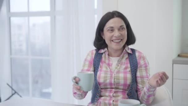 Porträtt av glad brunett Kaukasiska kvinna dricka te eller kaffe med vän och skrattar. Glada vuxna kvinnor tillbringar dagen tillsammans inomhus. Lycka, fritid, vänskap. — Stockvideo