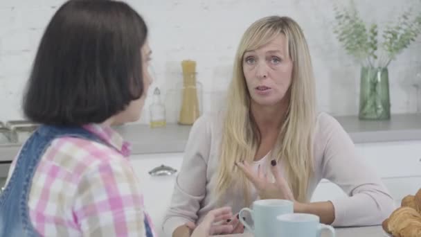 Portret van een volwassen blonde blanke vrouw die praat met een brunette vriendin en gebaart. Vrouwen roddelen als zittend aan tafel met koffie of thee en snacks. Vrije tijd, levensstijl, vriendschap. — Stockvideo