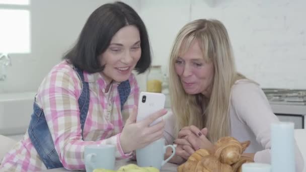 Primer plano de dos mujeres adultas caucásicas mirando la pantalla del teléfono inteligente y sonriendo. Mujeres amigas positivas descansando en el interior los fines de semana. Ocio, felicidad, amistad . — Vídeos de Stock