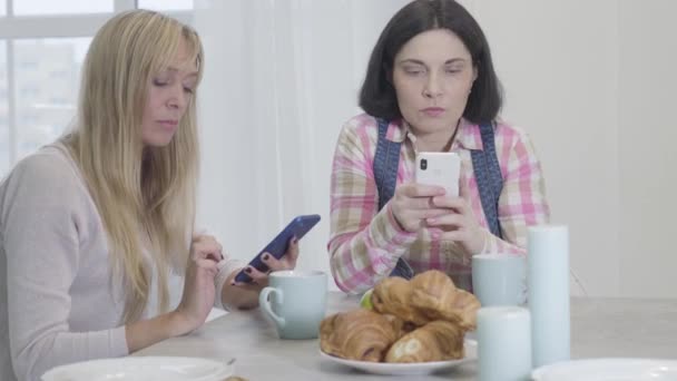 Retrato de duas mulheres caucasianas sérias sentadas à mesa e olhando para telas de smartphones. Amigos adultos do sexo feminino que se comunicam online enquanto descansam dentro de casa. Vício em Internet, redes sociais . — Vídeo de Stock