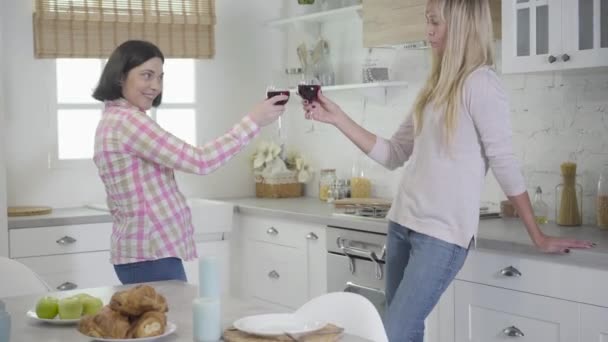 Két kaukázusi nő csörömpöl borosüvegben és iszik. Boldog női barátok pihennek otthon hétvégenként. Szabadidő, boldogság, életmód. — Stock videók