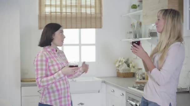 Vue latérale d'une femme caucasienne positive debout avec du verre à vin et parlant à un ami adulte blond. Des gens joyeux qui se reposent le week-end à la maison. Loisirs, bonheur, mode de vie . — Video