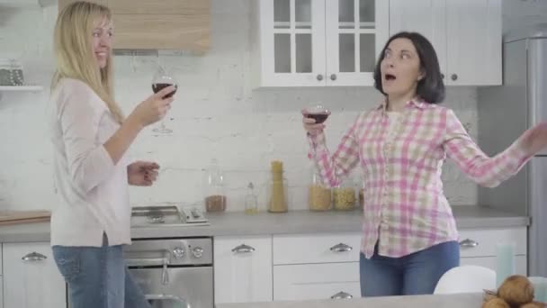 Femmes blanches adultes positives tenant des verres à vin et dansant dans la cuisine moderne. Deux amis qui s'amusent le week-end à la maison. Loisirs, bonheur, mode de vie . — Video