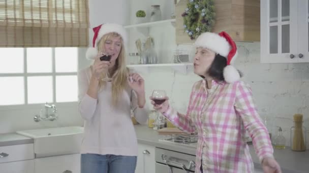 Двоє дорослих кавказьких жінок у червоних різдвяних капелюхах танцюють і співають як вино на сучасній кухні. Веселі друзі насолоджуються вихідними в приміщенні. Дружба, дозвілля, відпочинок. — стокове відео