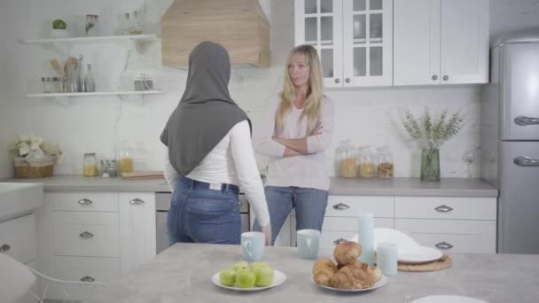Felnőtt szőke, kaukázusi nő portréja, amint muzulmán barátnőjével veszekszik. Két nő vitatkozik a modern konyhában. Kommunikációs problémák, félreértés. — Stock videók