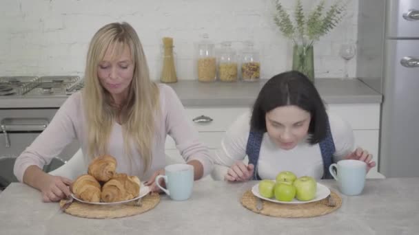 Brunette Caucasian woman smelling green apples as her blond friend taking croissant from the plate. Healthy and unhealthy eating, lifestyle. — Stock Video