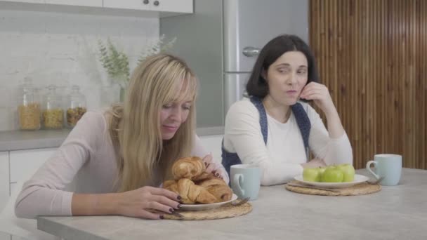 Plump Caucasian woman sitting in front of plate with apples and looking jealously at slim blond friend smelling tasty croissants. Healthy lifestyle, unhealthy food, body weight problems. — Stock Video