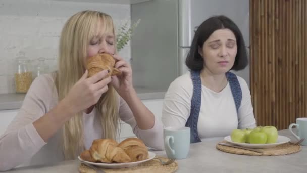 Retrato de mujer caucásica regordeta disgustada mirando celosamente a un delgado amigo rubio comiendo sabrosos croissants y mordiendo manzana. Estilo de vida saludable, alimentos poco saludables, problemas de peso corporal . — Vídeo de stock