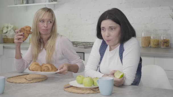 Upset plump Caucasian woman chewing apple, her slim friend giving her plate with tasty croissants, happy lady eating them hungrily. Healthy lifestyle, unhealthy food, body weight problems. — Stock Video