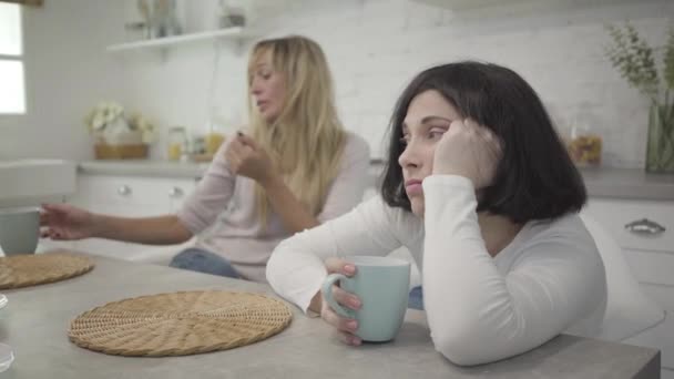 Primer plano de la morena adulta mujer caucásica sentada con taza de café en la mesa. Su amiga rubia bebiendo bebida caliente al fondo. Una dama molesta pasando el día con otro. Amistad, descanso . — Vídeos de Stock