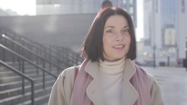 Ritratto di affascinante donna caucasica che guarda la macchina fotografica e sorride. Bella donna d'affari bruna in piedi alla luce del sole sulla strada della città prima dell'incontro. Stile di vita, felicità, successo . — Video Stock