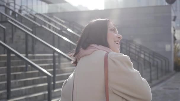 Brunette Kaukasische vrouw draait zich om naar camera en lacht. Positieve jongedame die buiten rust voor de vergadering. Lifestyle, geluk, succes. — Stockvideo