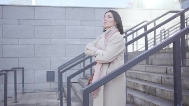 Side view of adult brunette Caucasian woman waiting for someone outdoors. Confident serious businesslady standing on stairs dressed in elegant coat. Meetings, business, lifestyle. — Stock Video