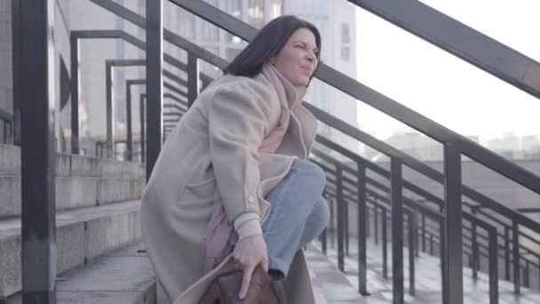 Pohled na mladou atraktivní bělošku sedící na schodech s bolestí kotníku. Mladá brunetka byznysmena trpí kvůli rychlému tempu života. Tvrdá práce, přepracování, zdravotní péče. — Stock video