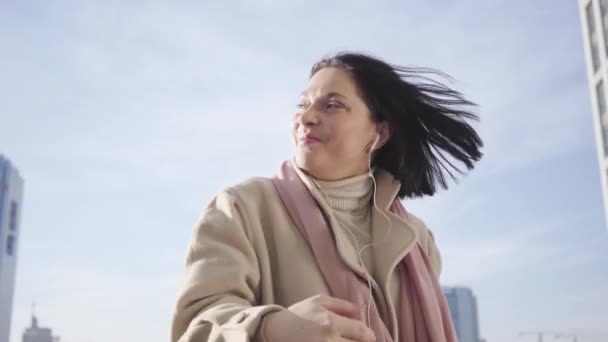 Närbild av ung vit kvinna i hörlurar stående på stadens gata och leende. Leende brunett affärerslady ha kul. Hobby, livsstil, vila. — Stockvideo