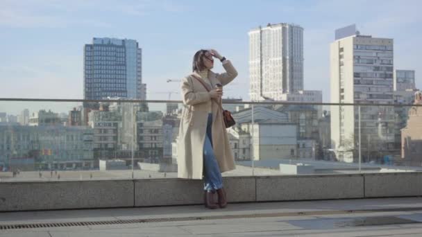 Young Caucasian businesswoman standing with coffee cup in the city and looking away. Confident brunette woman in elegant beige coat having break outdoors. Lifestyle, resting, elegance. — Stock Video