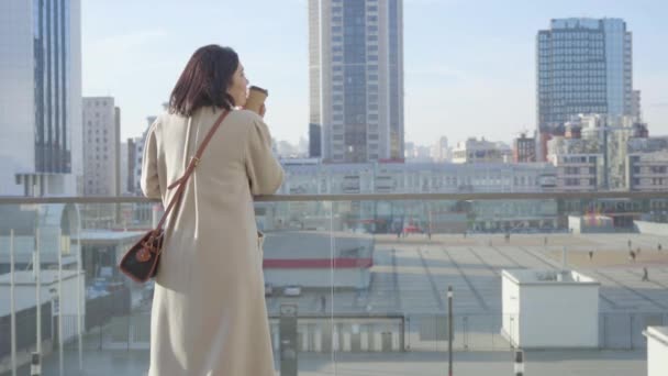 Back view of young Caucasian businesswoman standing with coffee cup and looking at city street. Confident woman in elegant beige coat having break outdoors. Lifestyle, resting, elegance. — Stock Video